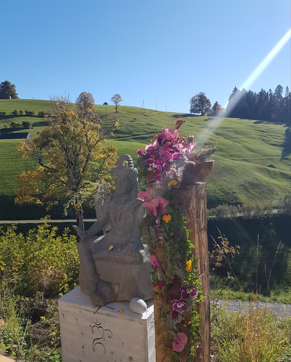 Steinerne Shiva-Figur mit OM-Symbol und Blumen vor den Toggenburger Hügeln im Karunahaus